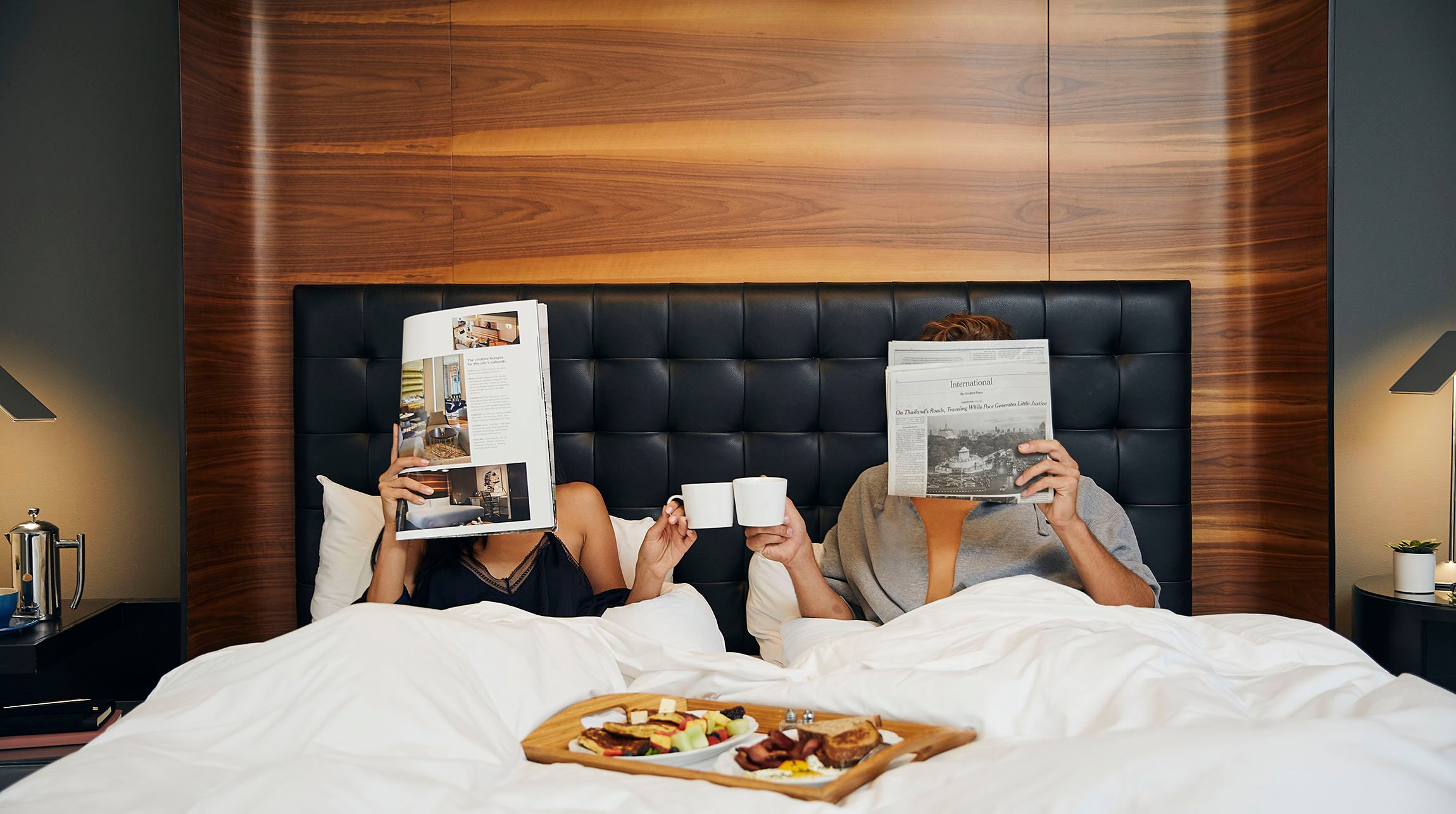 Couple in in Zetta Hotel bed, reading magazines, while having breakfast in bed.